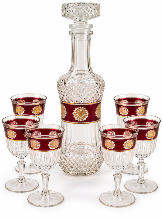 A clear decanter with a stopper, surrounded by six matching crystal goblets. Each goblet has a deep red band with a gold sunburst pattern near the rim, set against a plain white background.