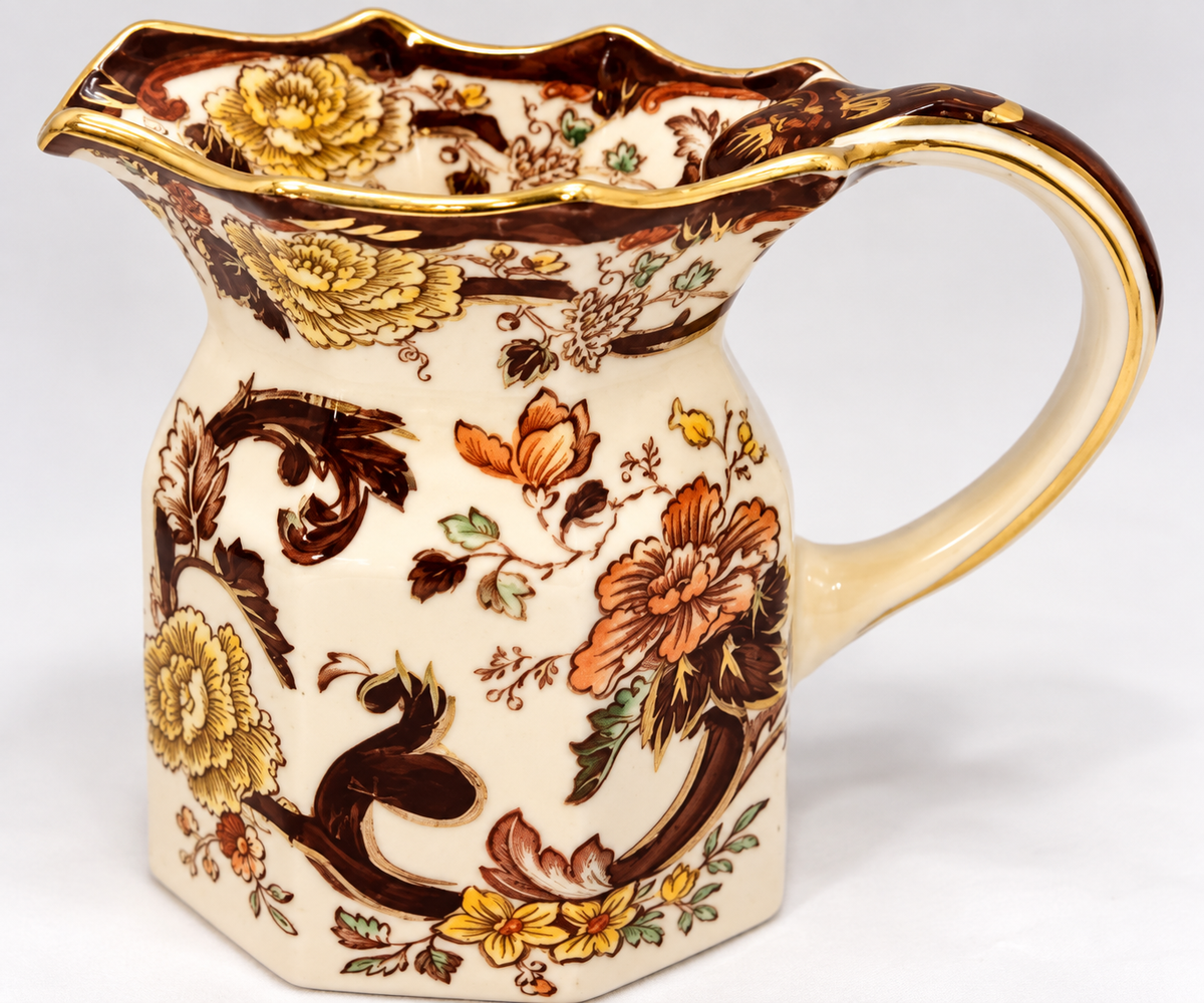 A vintage ceramic pitcher with a cream-colored glaze, decorated with brown floral and scroll motifs, yellow flowers, and gold trim on the rim and handle.